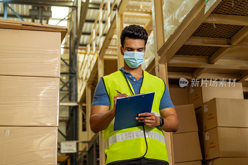 Young Indian factory warehouse worker wearing a protective face mask while working in logistic industry. 30s man checking item stock order during Covid 19 pandemic outbreak